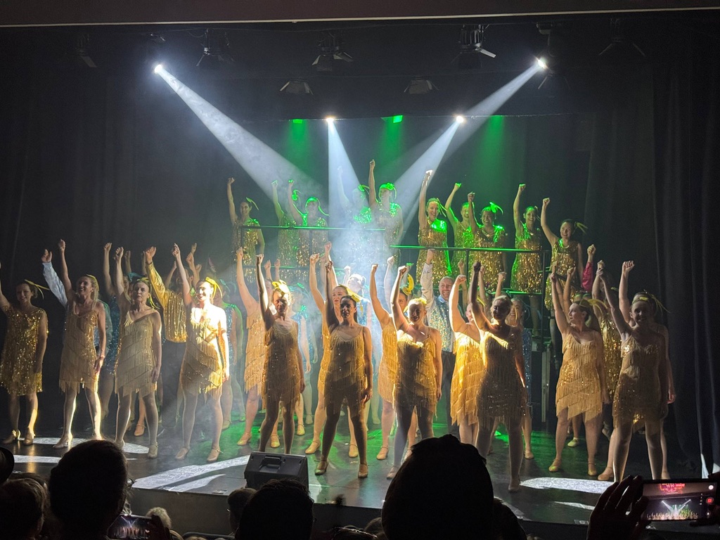 Ensemble cast under stage lights in gold flapper dresses
